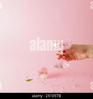 La main d'une femme tient un verre avec des pétales de rose, un glaçon est sur la table sur un fond rose Banque D'Images