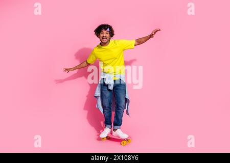 Photo de joyeux homme positif porter des vêtements jaunes à la mode équilibrant skateboard isolé sur fond de couleur rose Banque D'Images