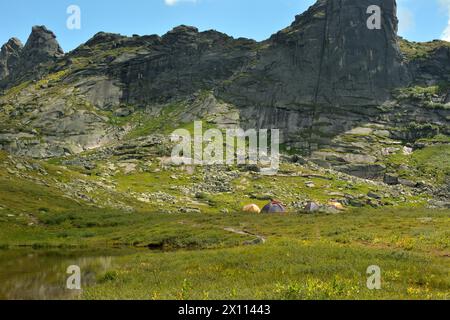 Plusieurs tentes touristiques sur la rive d'un petit lac au pied d'une haute falaise abrupte avec un pic pointu par une journée d'été ensoleillée. Parc naturel Ergaki, K Banque D'Images