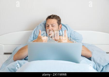 Jeune homme heureux communiquer par appel vidéo avec des amis montre le pouce vers le haut geste tout en étant allongé dans le lit et travailler sur un ordinateur portable à la maison. Joyeux Banque D'Images