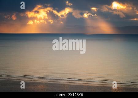 Ciel orageux et atmosphérique avec coucher de soleil doré regardant à travers la baie de Bideford depuis Saunton avec l'océan Atlantique calme, les rayons du soleil et la ligne de rivage. Banque D'Images