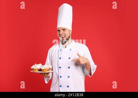 Heureux confiseur professionnel en uniforme pointant à de délicieux cupcakes sur fond rouge Banque D'Images