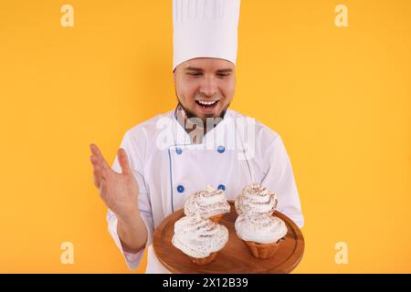 Heureux confiseur professionnel en uniforme tenant de délicieux cupcakes sur fond jaune Banque D'Images