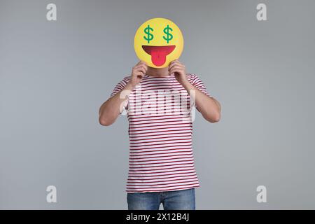 Homme tenant une émoticône avec des signes dollar au lieu des yeux sur fond gris Banque D'Images