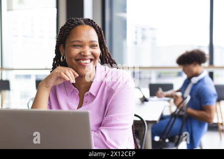 Jeune femme biraciale avec des cheveux tressés travaillant sur ordinateur portable, souriante Banque D'Images