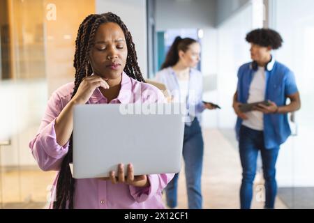 Femme biraciale tenant un ordinateur portable, pensant, collègues marchant derrière dans un bureau d'affaires moderne Banque D'Images