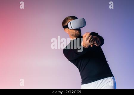 Un homme dans un casque VR balançant une batte de baseball tout en bandant les yeux dans un cadre de studio, mettant en vedette un entraînement sportif en réalité virtuelle. Banque D'Images