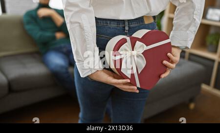 Une femme surprend un homme avec un cadeau en forme de cœur dans un salon confortable, incarnant l'amour et la convivialité. Banque D'Images