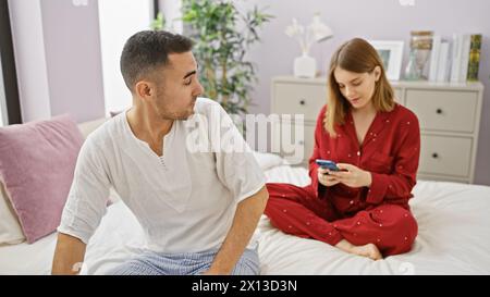 Un couple en pyjama se détend dans une chambre, la femme engrossée dans son téléphone et l'homme regardant. Banque D'Images