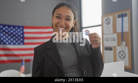 Une femme hispanique souriante tenant un autocollant «voté», avec le drapeau américain derrière elle dans un cadre intérieur. Banque D'Images