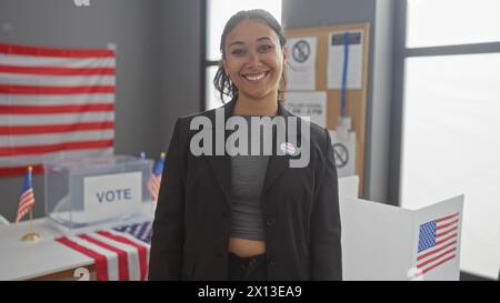 Jeune femme hispanique souriante avec l'autocollant "j'ai voté" au bureau de vote américain avec des drapeaux Banque D'Images