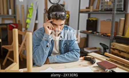 Un homme hispanique réfléchi avec une barbe portant des lunettes de sécurité se reflète dans un atelier de menuiserie encombré Banque D'Images