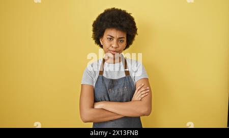 Femme africaine confiante avec les bras croisés debout sur un fond jaune portant un tablier en denim Banque D'Images