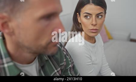 Un couple hispanique apparaît contemplatif dans leur salon, la femme paraissant anxieuse aux côtés de son homme sur le canapé. Banque D'Images