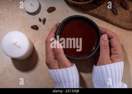 Boisson cérémonielle au cacao. Chocolat cérémonial chaud dans une tasse noire avec des fèves de cacao. Mains de femme tenant une tasse de cacao. Préparation de boisson au chocolat biologique saine Banque D'Images