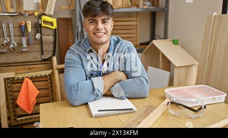 Attrayant jeune homme hispanique, un charpentier professionnel, assis les bras croisés, souriant au travail dans son atelier animé de menuiserie. Banque D'Images