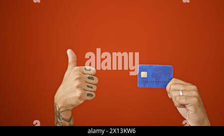 Les mains tatouées de l'homme poussent une carte de crédit sur un fond rouge isolé avec un geste correct - les pouces vers le haut en approbation Banque D'Images