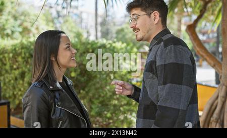 Un couple heureux s'engage dans une conversation animée dans une rue urbaine ensoleillée, entourée de verdure et de vie urbaine. Banque D'Images