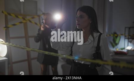 Deux enquêteurs femmes analysant une scène de crime à l'intérieur avec une lampe de poche, un ruban de mise en garde et des preuves. Banque D'Images