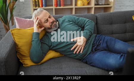 Un homme barbu d'âge moyen allongé sur un canapé à la maison, semblant souffrir de douleurs à l'estomac. Banque D'Images