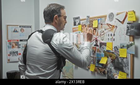 Un homme mûr analyse un tableau de preuve du crime dans le bureau d'un détective, entouré de photos, de notes et d'empreintes digitales. Banque D'Images