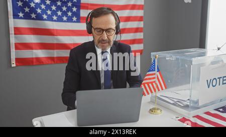 Un homme mûr portant des écouteurs est assis devant un ordinateur portable dans un centre de vote avec un drapeau américain et une urne. Banque D'Images