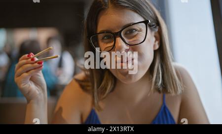 Aventure gastronomique inoubliable, belle femme hispanique mange joyeusement avec des baguettes dans l'intérieur moderne du restaurant asiatique Banque D'Images