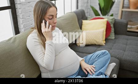 Une jeune femme enceinte est assise confortablement sur un canapé à la maison, engagée dans une conversation téléphonique. Banque D'Images