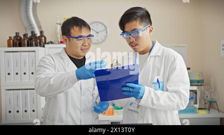 Deux hommes en blouse de laboratoire collaborent dans un laboratoire scientifique, discutant sur une planche à pince. Banque D'Images