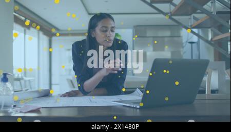 Image de taches jaunes flottant sur une femme biraciale travaillant sur un plan et utilisant un ordinateur portable au bureau Banque D'Images