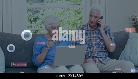 Image d'icônes de médias sociaux au-dessus d'un couple de personnes âgées caucasiennes utilisant des appareils électroniques à la maison Banque D'Images