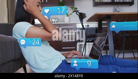 Image des icônes de médias sociaux et des numéros sur la femme biraciale à l'aide d'un ordinateur portable. Concept mondial de médias sociaux et de traitement des données image générée numériquement. Banque D'Images