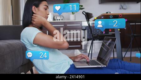 Image des icônes de médias sociaux et des numéros sur la femme biraciale à l'aide d'un ordinateur portable Banque D'Images