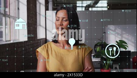 Image de chiffres et d'icônes changeants sur la femme biraciale debout bras croisés dans le bureau Banque D'Images