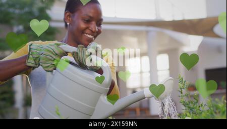 Image de coeurs verts sur la femme afro-américaine heureuse arrosant des plantes Banque D'Images