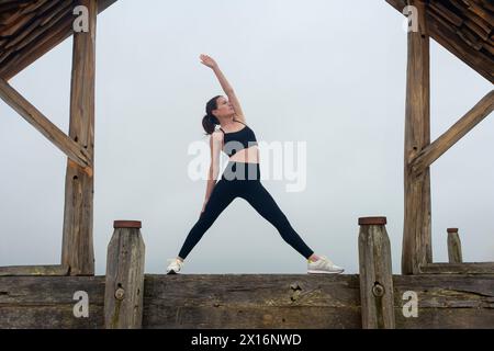Femme sportive faisant une pose de yoga guerrier inversée, à l'extérieur par un bâtiment en bois Banque D'Images