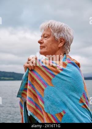 Un nageur en eau libre senior se réchauffe avec une serviette après une baignade rafraîchissante dans le lac d'eau froide - Woman Swim Outdoors Health UK Banque D'Images