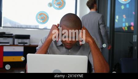 Image de plusieurs icônes de graphique sur l'homme afro-américain fatigué regardant l'ordinateur portable dans le bureau Banque D'Images