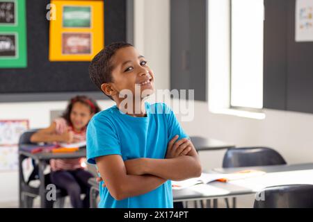 À l'école, jeune garçon biracial debout, les bras croisés, regardant réfléchi dans la salle de classe Banque D'Images