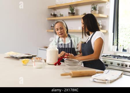 La grand-mère asiatique et la petite-fille adolescente biraciale cuisent ensemble à la maison. Les deux portant des tabliers, la femme âgée a les cheveux gris, le jeune a les cheveux foncés Banque D'Images