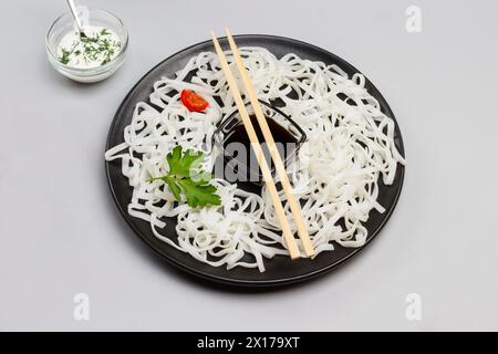 Nouilles de riz avec sauce, bâtonnets de bambou sur assiette noire. Tomates cerises dans un bol avec sauce blanche sur la table. Fond gris. Vue de dessus. Banque D'Images