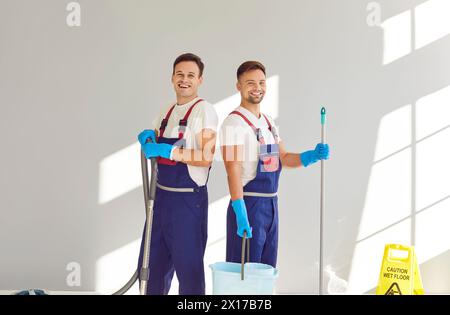 Portrait de deux concierges masculins heureux et souriants en uniformes avec du matériel de nettoyage Banque D'Images