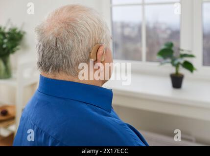 Vue arrière d'un homme âgé aux cheveux gris portant une prothèse auditive moderne dans son oreille à la maison. Banque D'Images