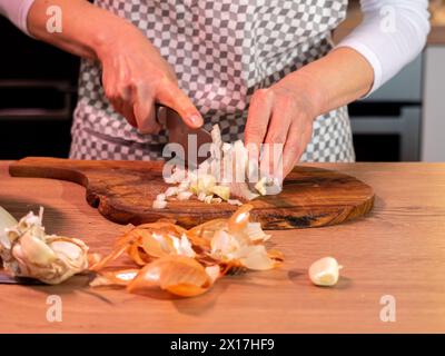 couper l'oignon sur une planche de bois, cuisiner dans un tablier à carreaux avec un couteau à la main, dessus de table en bois, photo de devant Banque D'Images