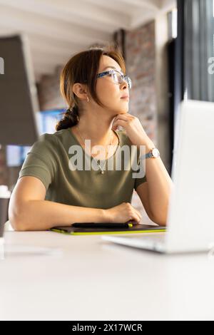 Femme asiatique d'âge moyen portant des lunettes regardant l'écran d'ordinateur portable dans un bureau d'affaires moderne Banque D'Images