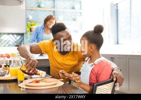 Père afro-américain servant le petit déjeuner à sa fille à la maison, mère en arrière-plan Banque D'Images