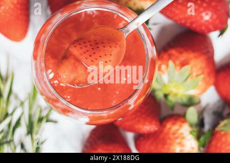 Gros plan d'une cuillère ramasse une confiture de fraises fraîche dans un pot en verre. Il y a plusieurs fraises sur la table Banque D'Images