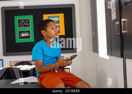 À l'école, en classe, jeune garçon biracial tenant un smartphone, le regardant Banque D'Images