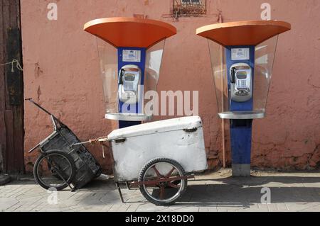 Marrakech, deux téléphones publics orange et blanc sur un mur rouge, à côté d'un vieux chariot, Marrakech, Maroc Banque D'Images