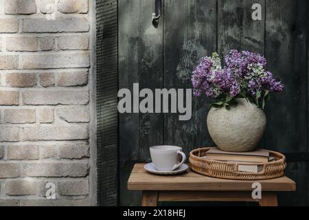 Petit déjeuner de printemps ferme nature morte. Bouquet de fleurs de lilas blanc violet dans un vase texturé avec tasse de café. Plateau en osier, livres. Vieille porte en bois floue Banque D'Images